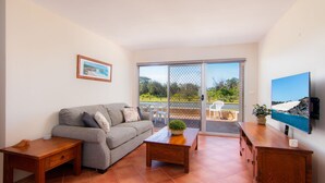 En stue med en grå sofa, et træbord og et tv-skab. Der er en skydedør, som fører ud til en udendørs terrasse med et bord og stole.