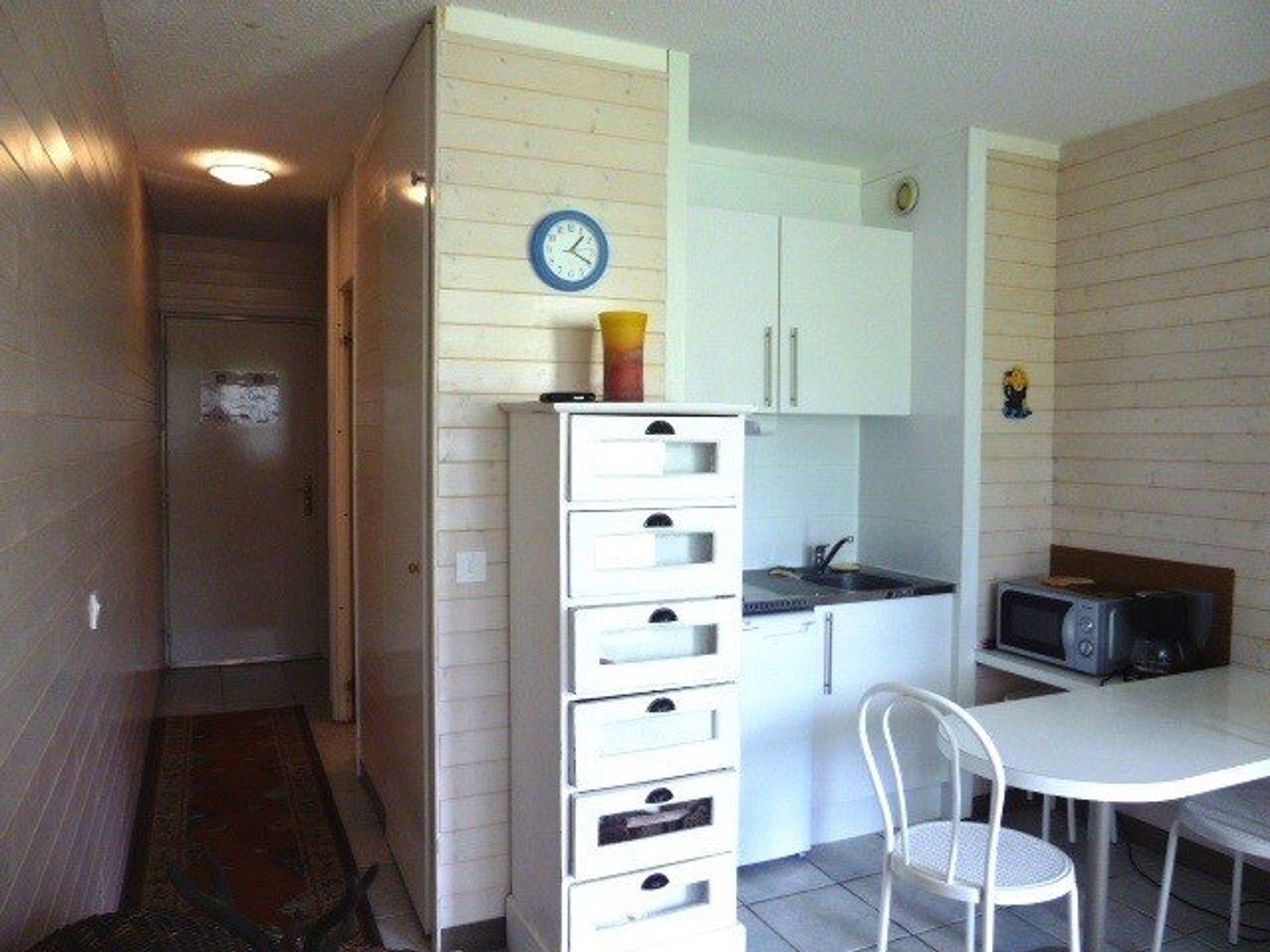 A compact kitchen with white cabinets, a microwave, a clock, and a small dining table with a chair.