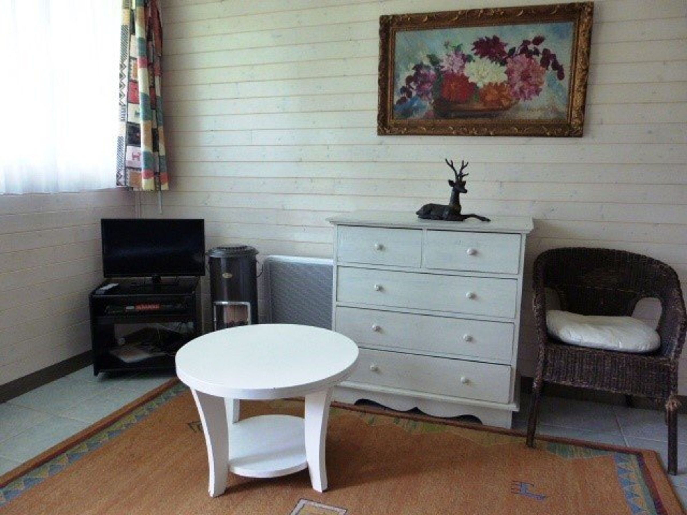 A room with a white table, a wicker chair, a dresser, a television, and a painting on the wall.
