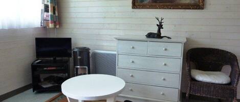 A room with a white table, a wicker chair, a dresser, a television, and a painting on the wall.
