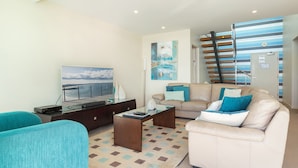A modern living room with a sofa, coffee table, and a flat-screen TV. There is a staircase with a railing leading to an upper level.