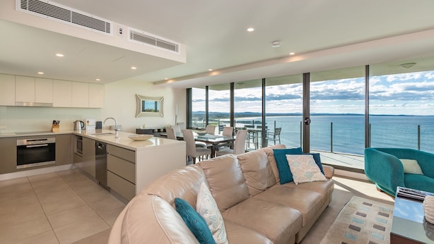 A modern kitchen with stainless steel appliances, a dining area with a view of the sea, and a cozy seating area with a leather sofa and teal armchair.