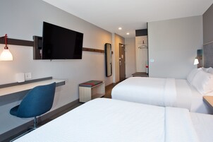 Habitación de hotel con dos camas, un televisor montado en la pared, un escritorio con una silla y un espejo.