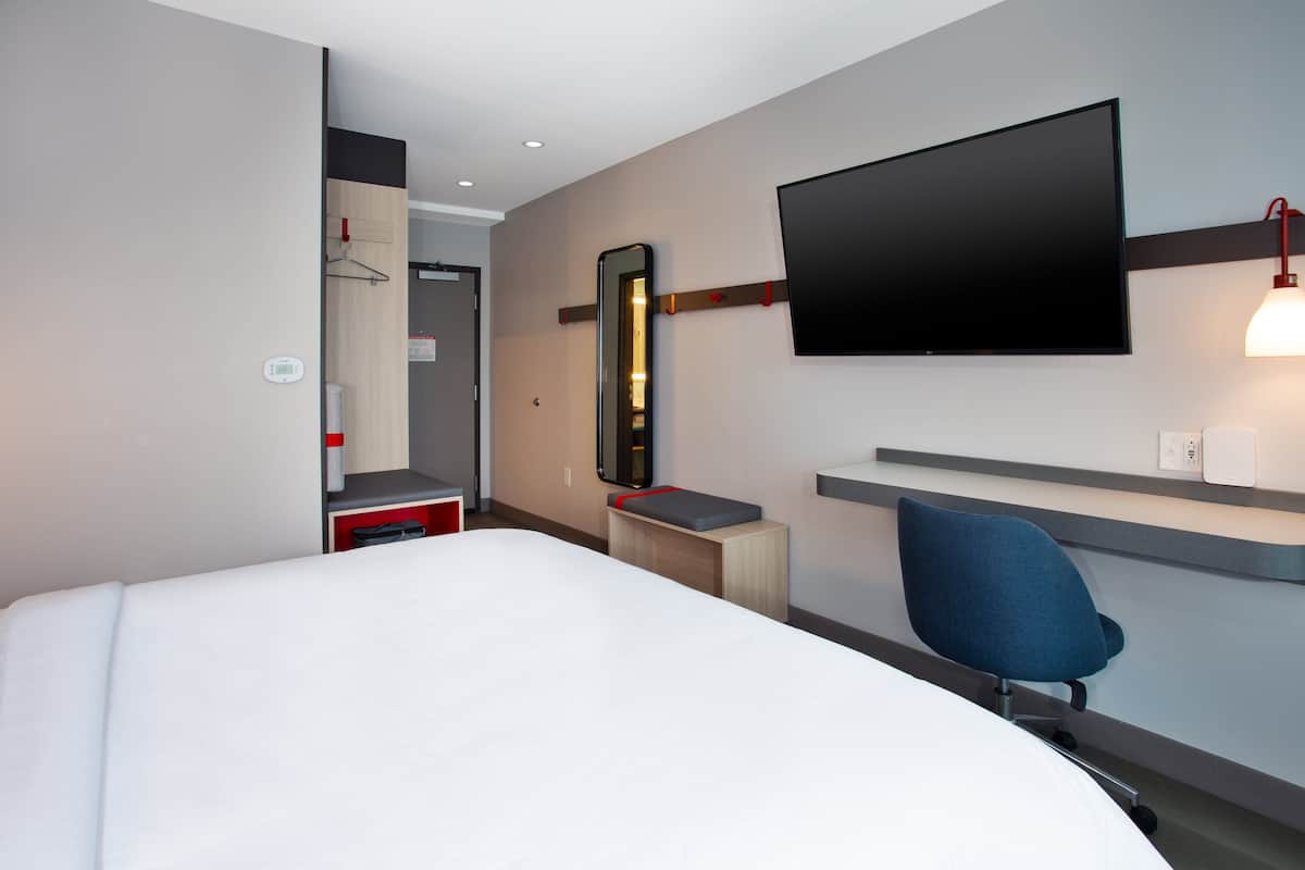A hotel room with a bed, a desk, a chair, a TV mounted on the wall, and a mirror.