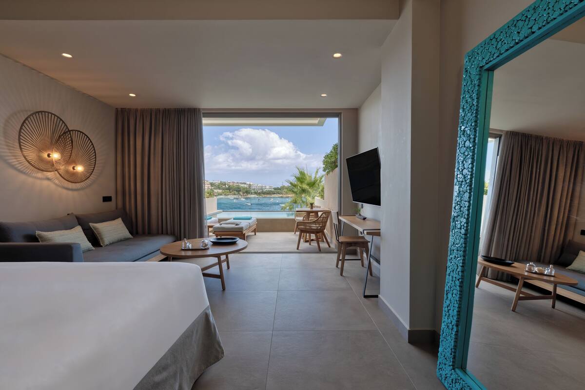 A modern hotel room with a large bed, a seating area with a table and chairs, a TV, and a balcony with a view of the sea.