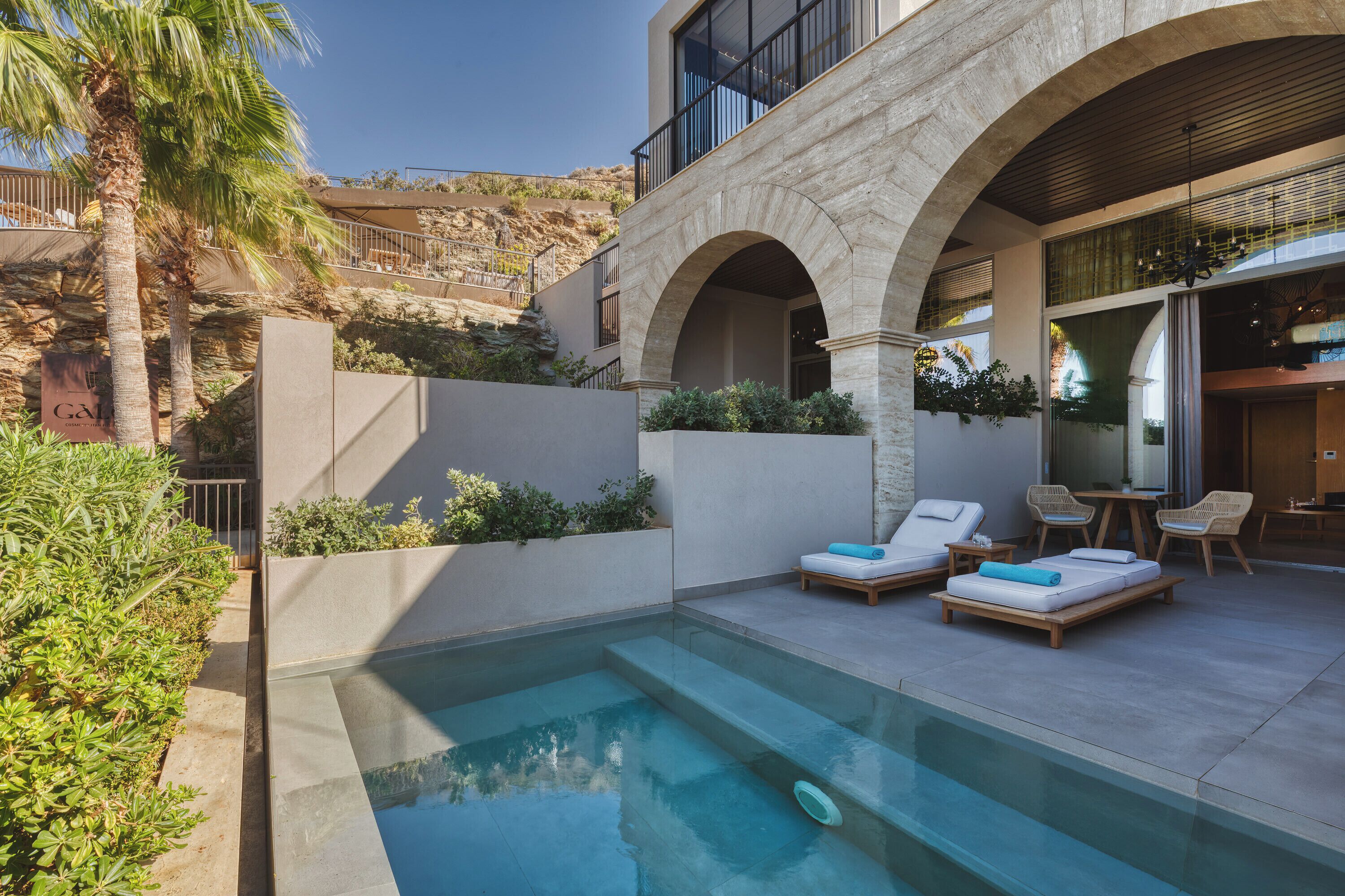 Zona de piscina con sillones de descanso y una mesa, rodeada por un muro de piedra y vegetación.