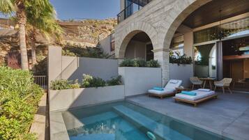 Zona de piscina con sillones de descanso y una mesa, rodeada por un muro de piedra y vegetación.