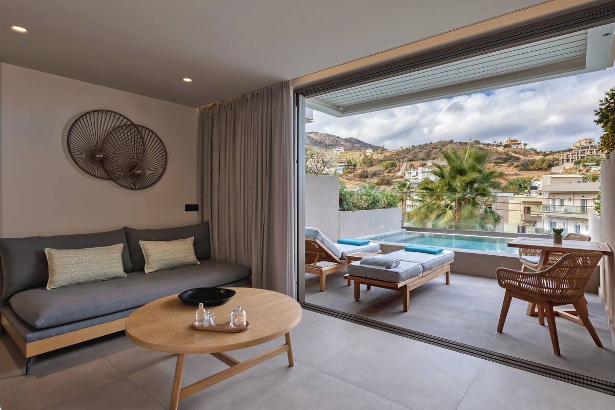 A modern living room with a sofa, a round table, and a view of a pool and hillside.