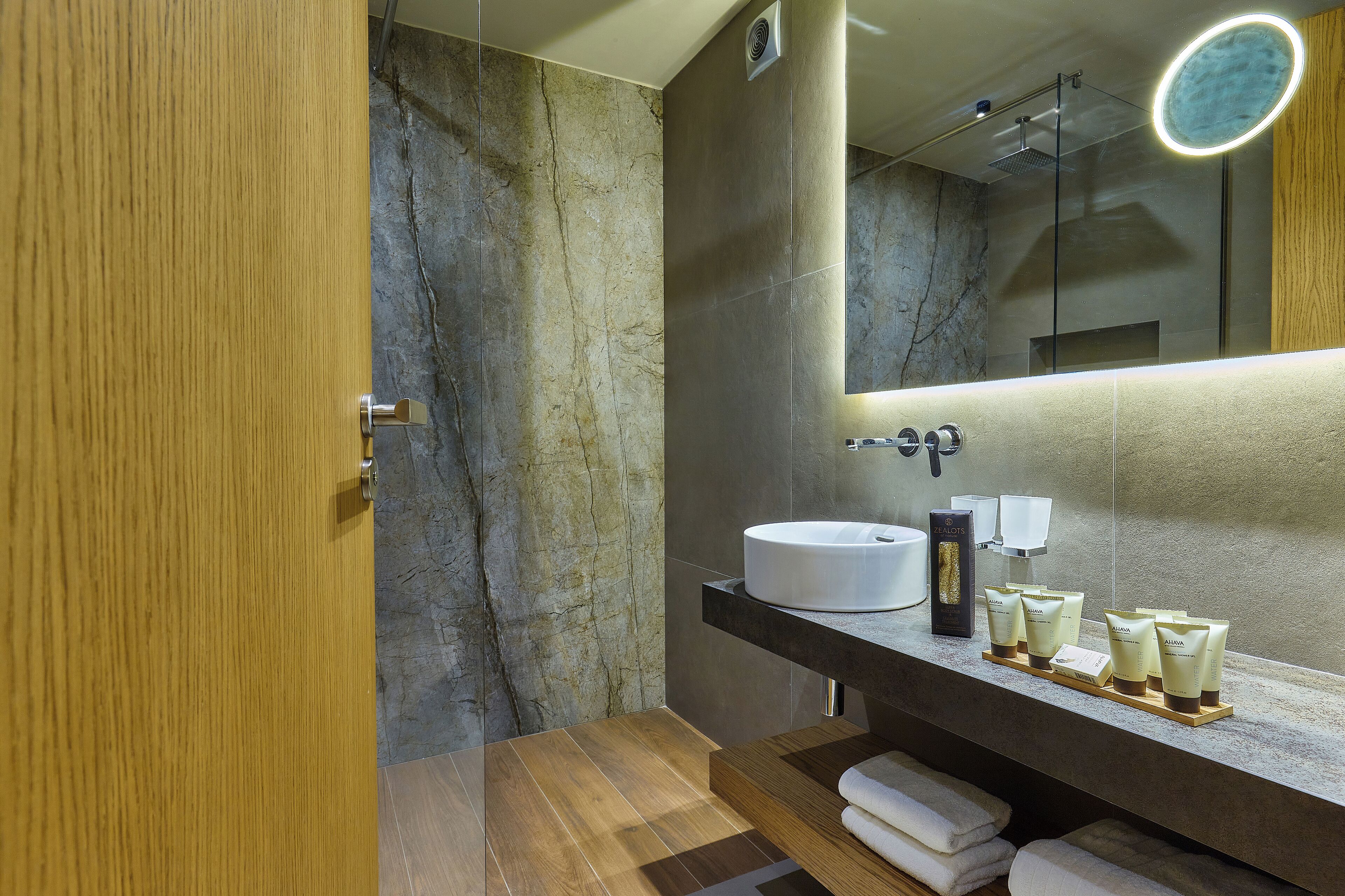 Een moderne badkamer met een marmeren wand, een houten deur, een ronde spiegel en een witte wasbak.
