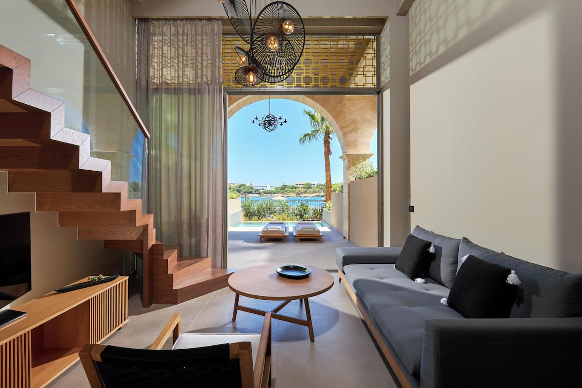 A modern living room with a staircase, a sofa, a round table, and a view of a pool and palm trees.