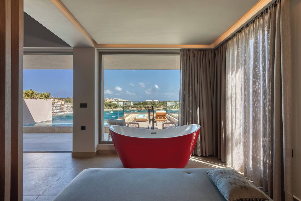 A modern hotel room with a large window, a red bathtub, and a view of a pool and buildings.