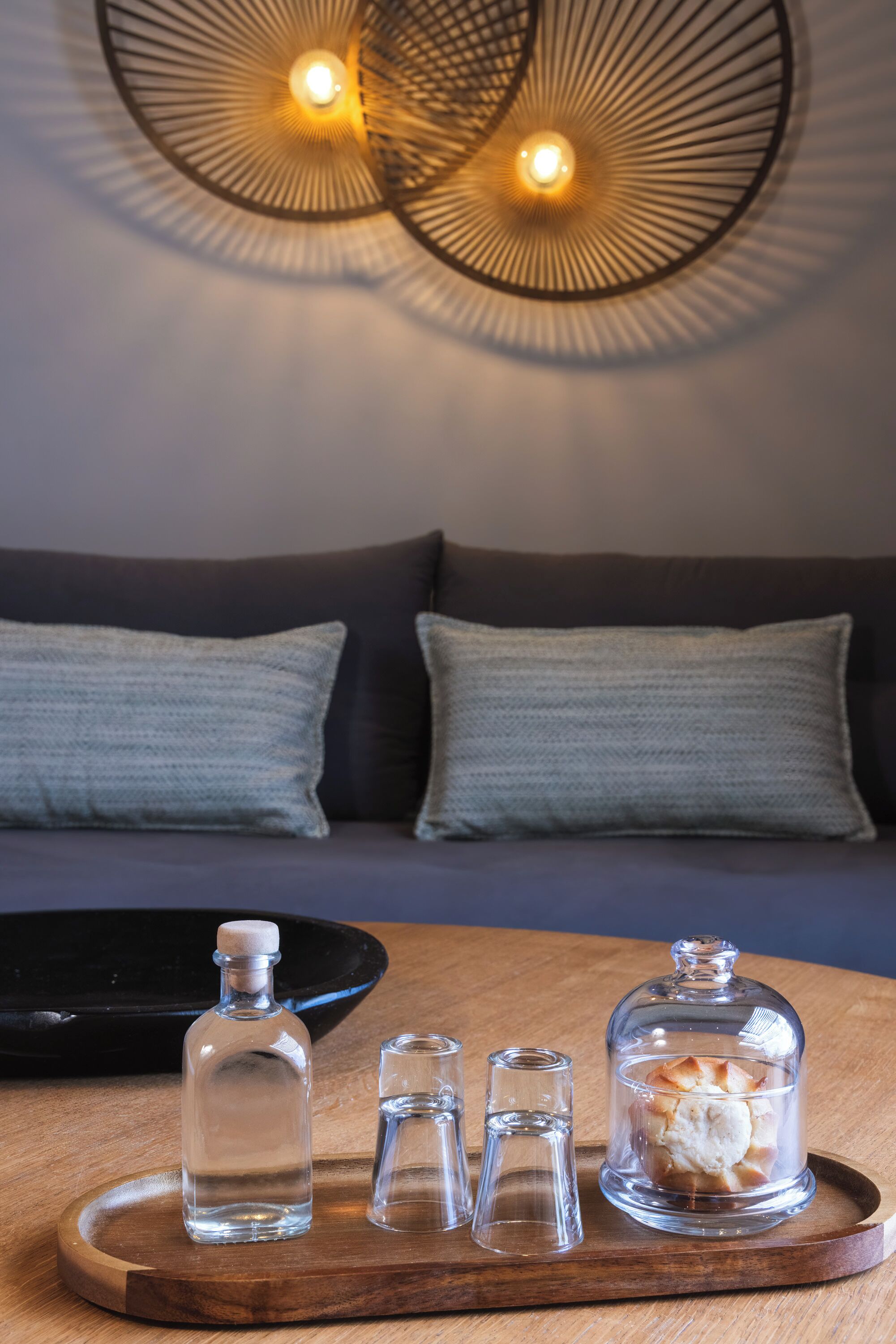 A wooden tray with three glass bottles and a glass dome containing a pastry on a wooden table in a room with a dark sofa and patterned wall decor.