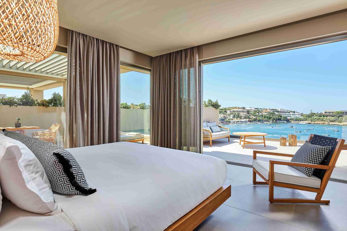 A modern hotel room with a large bed, a balcony with a view of the sea, and a seating area with a table and chairs.