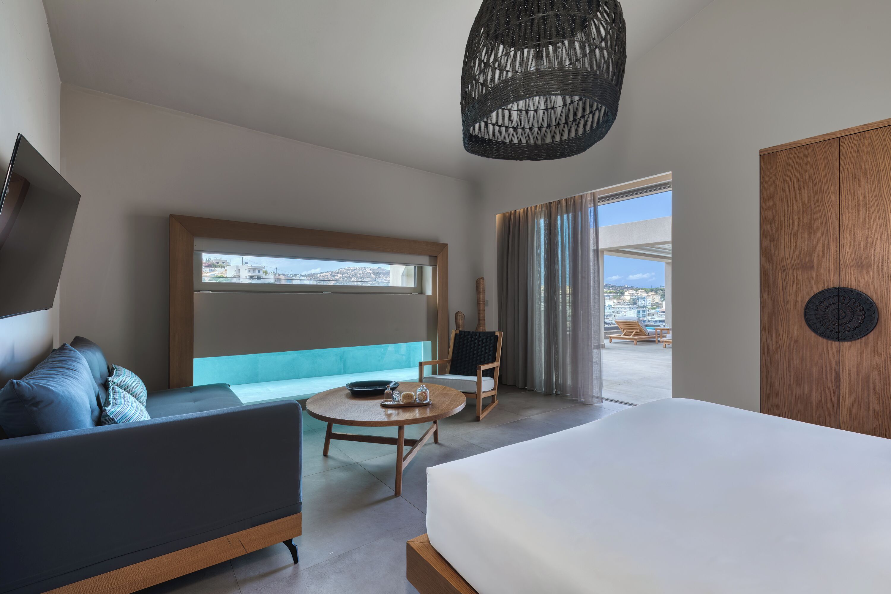 A modern hotel room with a bed, a sofa, a round table, and a chair. There is a large window with a view of the city and a door with a circular design.