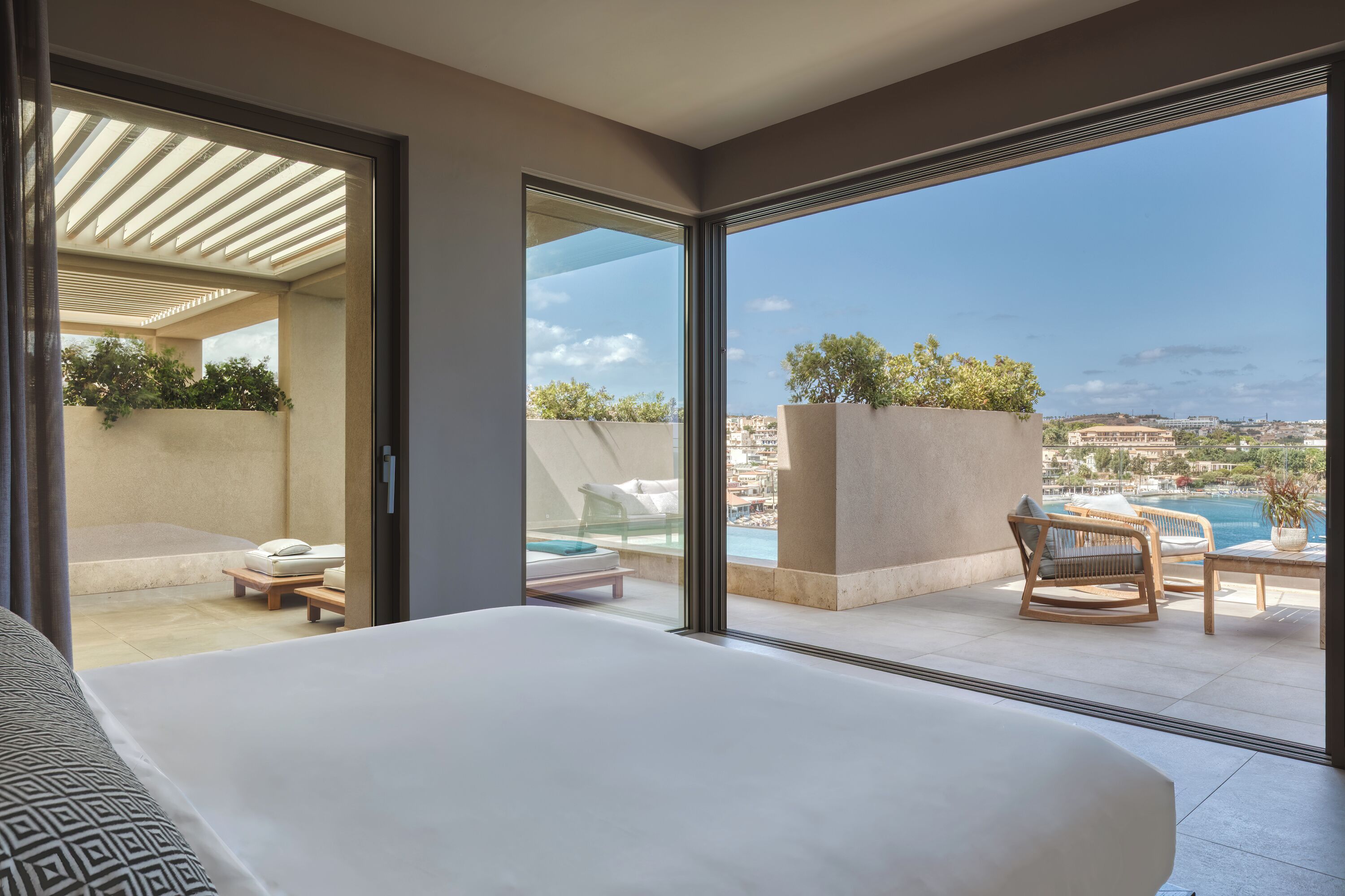 Habitación de hotel con un balcón grande, una cama, zona de estar con mesa y vistas a una piscina y al paisaje urbano.