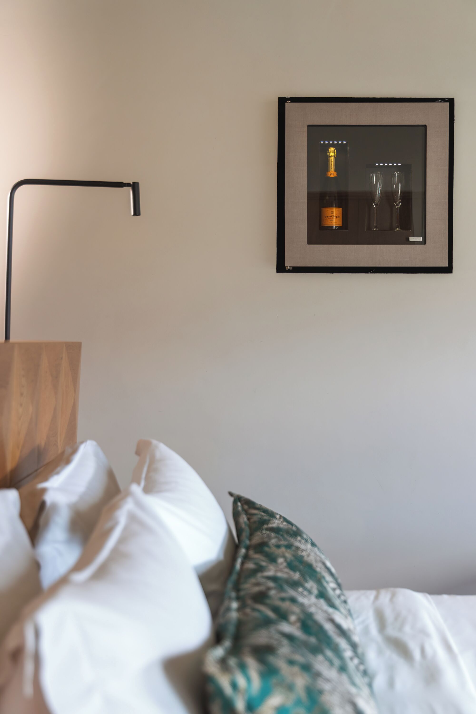 Una cama bien hecha con almohadas blancas y estampadas, un cuadro con una botella y copas en la pared, y una lámpara de pie moderna.