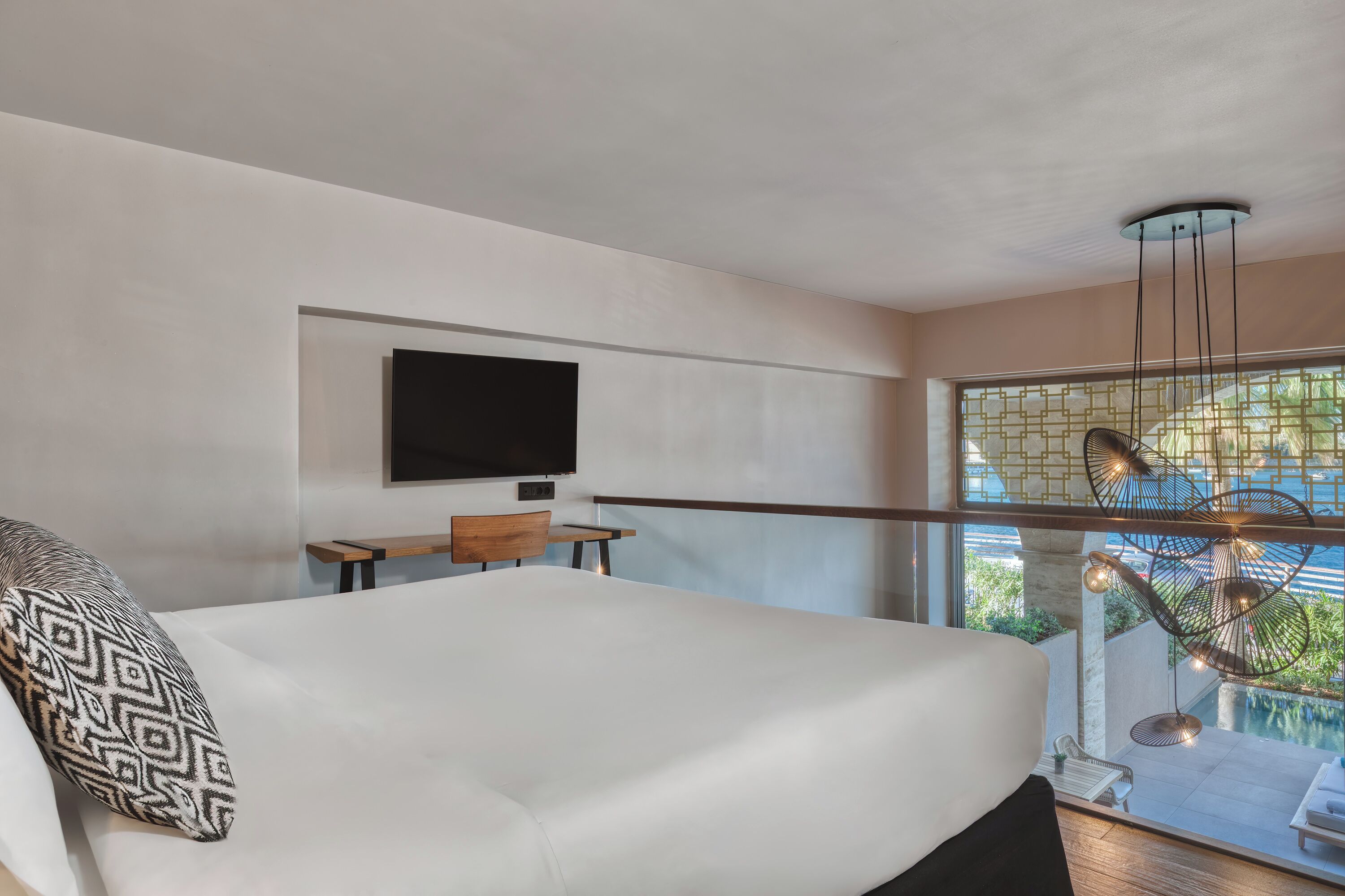 A modern hotel room with a large bed, a desk, a television, and a view of an outdoor pool area.