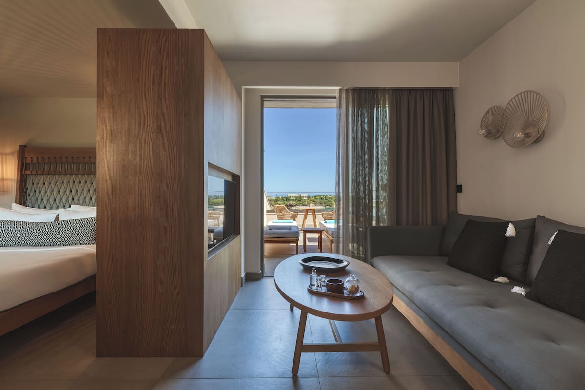A modern hotel room with a bed, a sofa, a dining table, and a balcony with a view.
