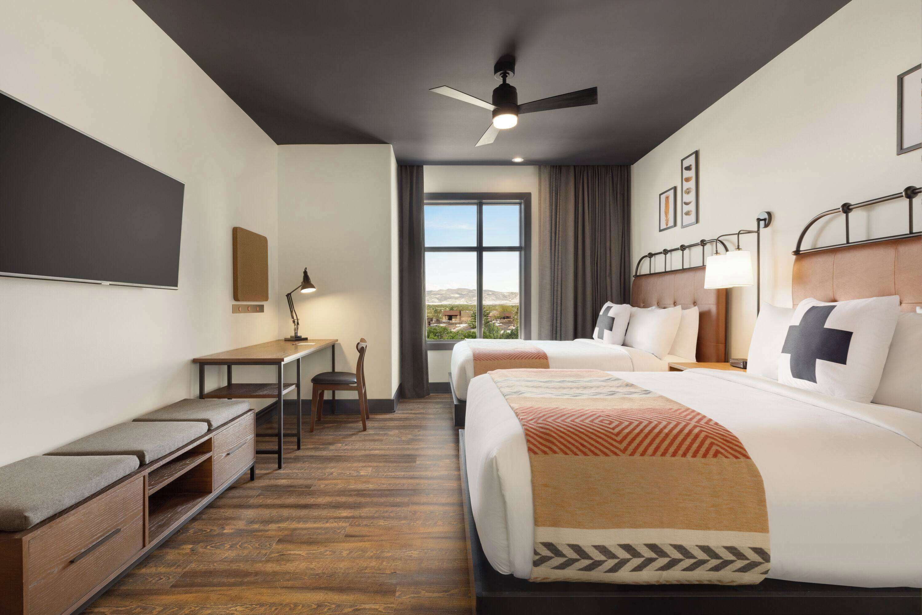 A hotel room with two beds, a desk, a chair, a ceiling fan, and a large window offering a view.