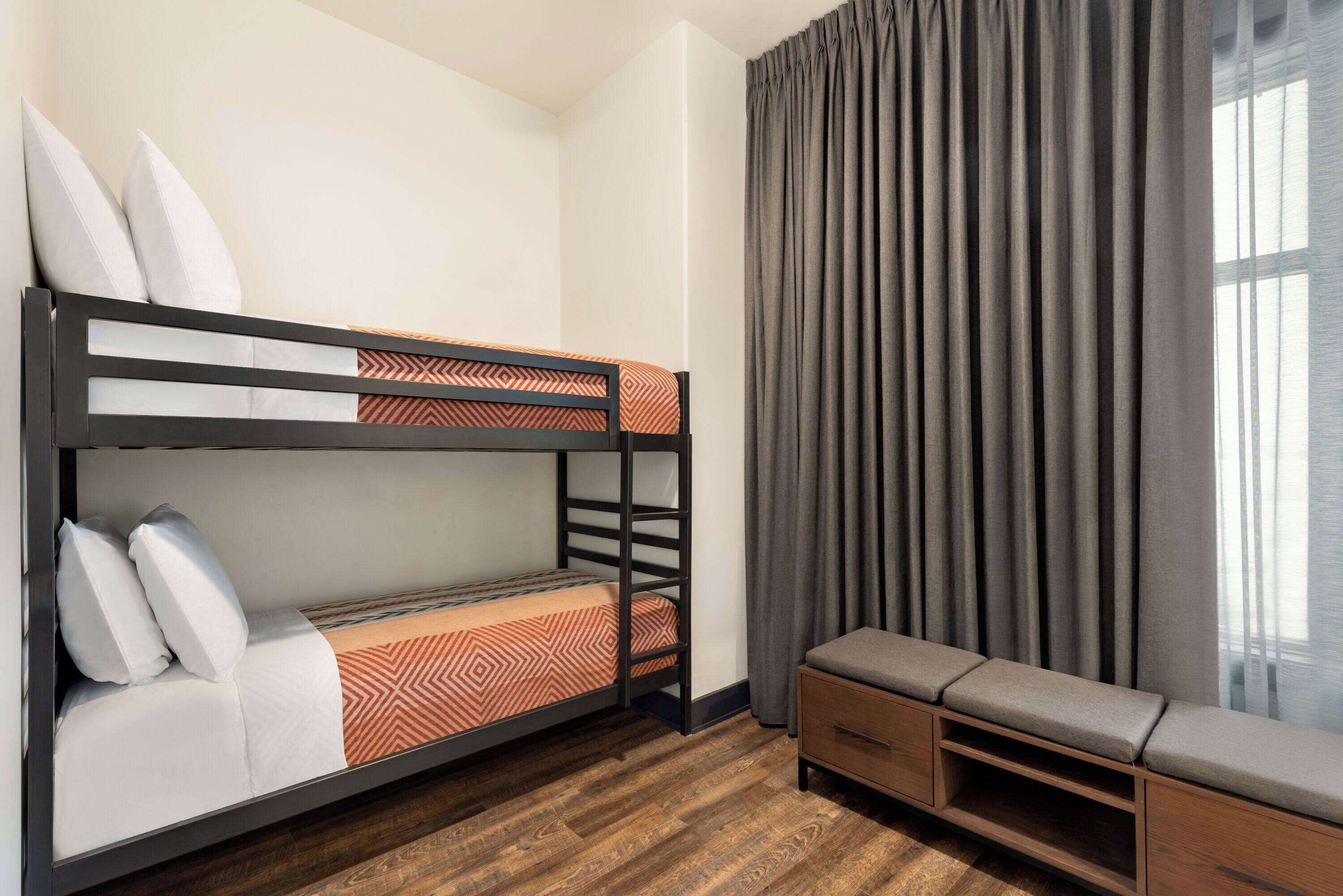 A bunk bed with a patterned bedspread, a wooden bench with storage, and a window with curtains.