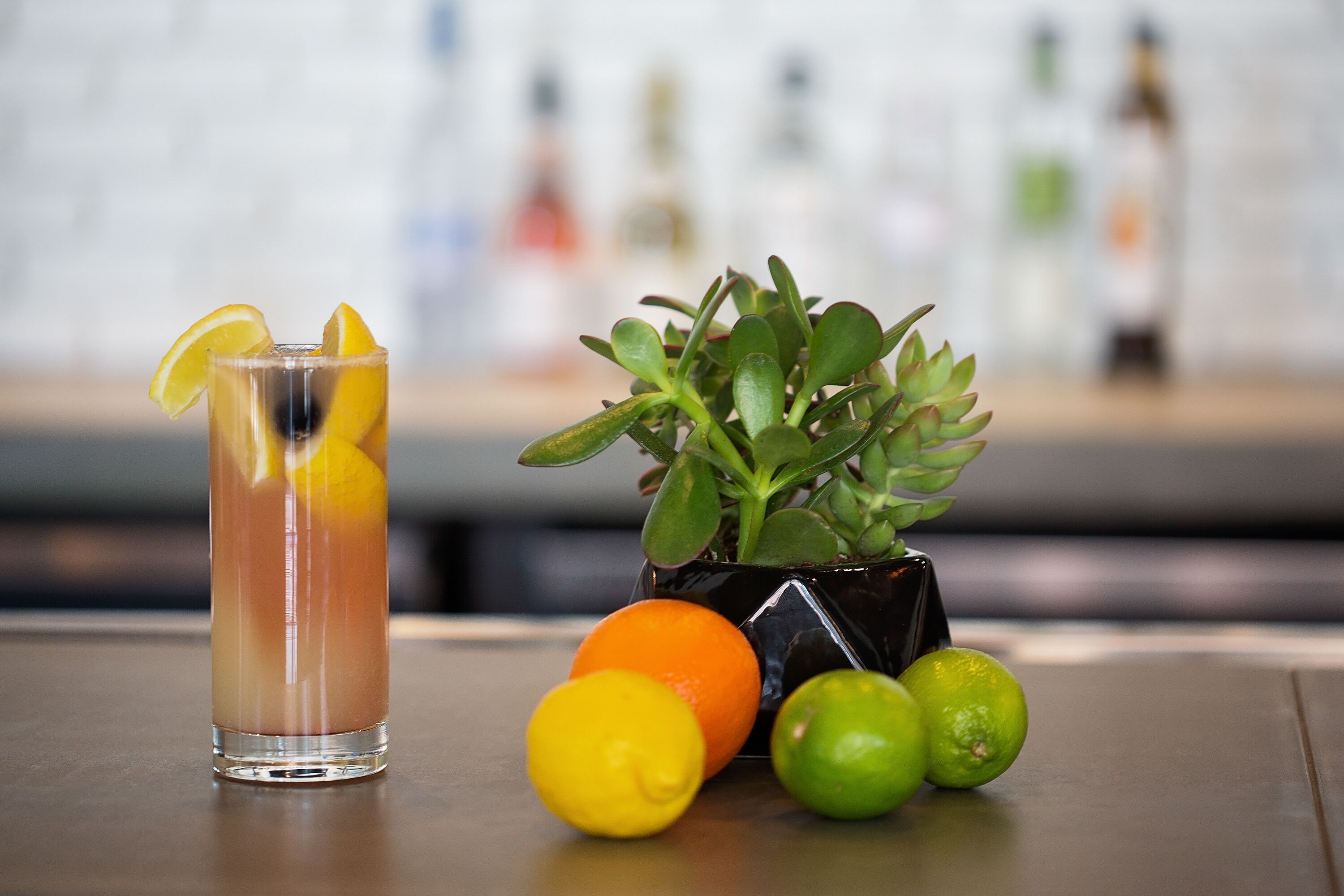 A clear glass with a beverage, a potted plant, and citrus fruits on a table.