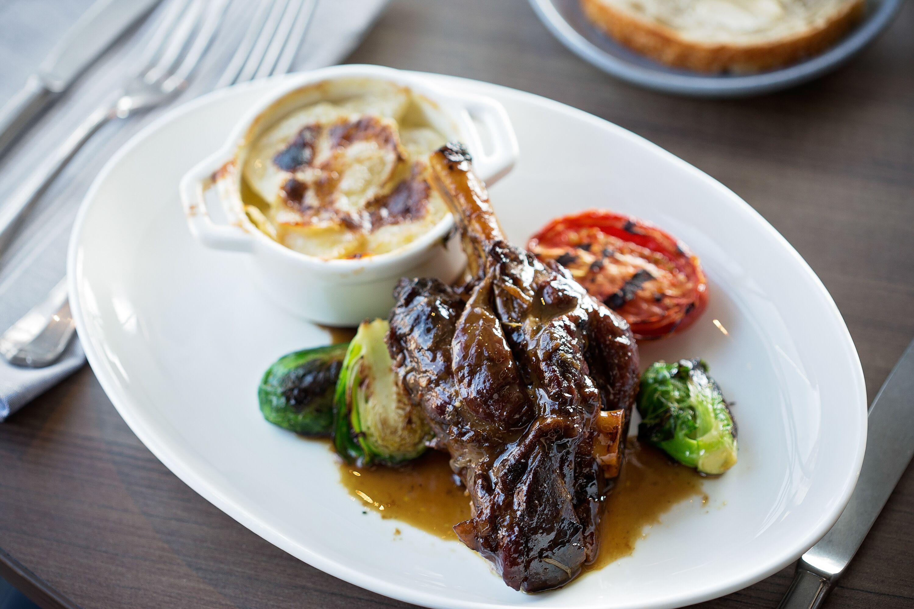 A plate of food with a lamb shank, roasted vegetables, and a side of mashed potatoes.