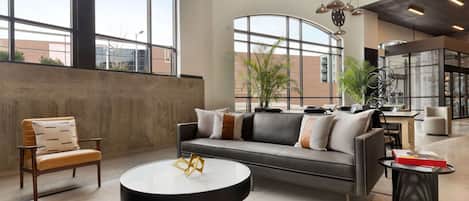 Un espacio lounge moderno con sofá, sillón y mesa redonda, ventanas amplias y lámparas colgantes.
