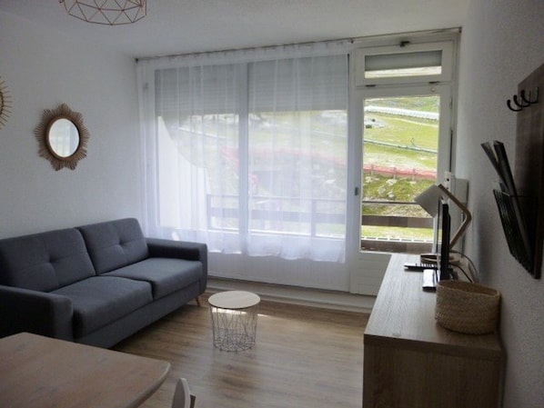 A modern living room with a grey sofa, a round coffee table, and a dining table. A large window with sheer curtains offers a view of a garden and a building.