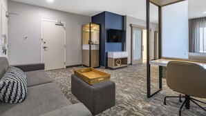 Ett modernt hotellrum med en grå soffa, ett bord i trä, ett skrivbord med en stol, en tv som är monterad på väggen och en stor spegel.