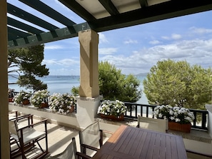 A covered outdoor seating area with wooden chairs and tables, potted white flowers, and a view of the sea with boats.