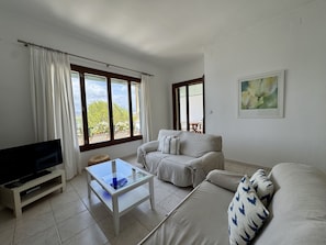 A living room with a sofa, a glass coffee table, a TV on a stand, and large windows with curtains.