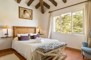 A bedroom with a wooden headboard, a bed with white bedding and a zebra-patterned pillow, two bedside lamps, a painting above the bed, a window with curtains, and a blue armchair. - Elegant 4 bedroom villa in Mancor de la Vall (Mancor de la Vall)