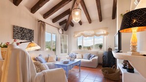 Un salon chaleureux avec un canapé, une table basse et une cheminée. Il y a des fenêtres avec des rideaux, un lustre et une vue sur l’extérieur.