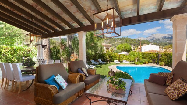 Un espace extérieur aménagé, abrité et doté de mobilier en osier, d’une table en verre, et offrant une vue sur une piscine et des montagnes.