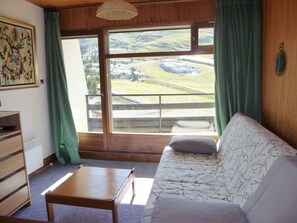Quarto com sofá, uma pequena mesa de madeira, um quadro e vista para uma paisagem através de uma grande janela.