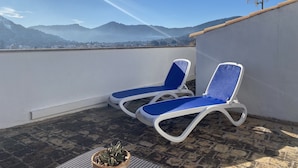 A rooftop terrace with two blue lounge chairs, a potted plant, and a view of distant mountains.