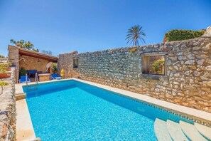 A stone-walled pool area with a clear blue pool, a small window, and a palm tree in the background. - Charming stone clad family villa with pool La Font (Pollensa / Pollença)