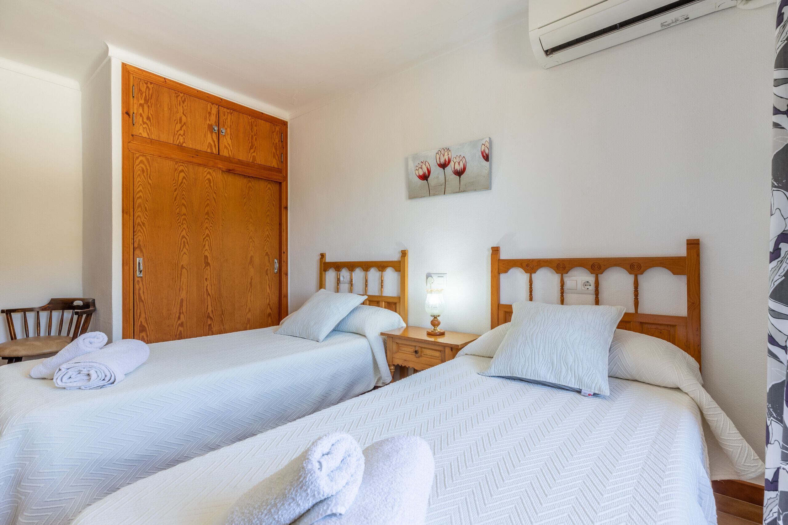 A hotel room with two single beds, a wooden wardrobe, a small table, a wall-mounted lamp, and a painting of flowers.