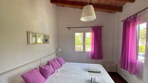 Une chambre à coucher avec un lit, des oreillers violets, un couvre-lit blanc, une lampe suspendue, une fenêtre avec des rideaux et un tableau accroché au mur.