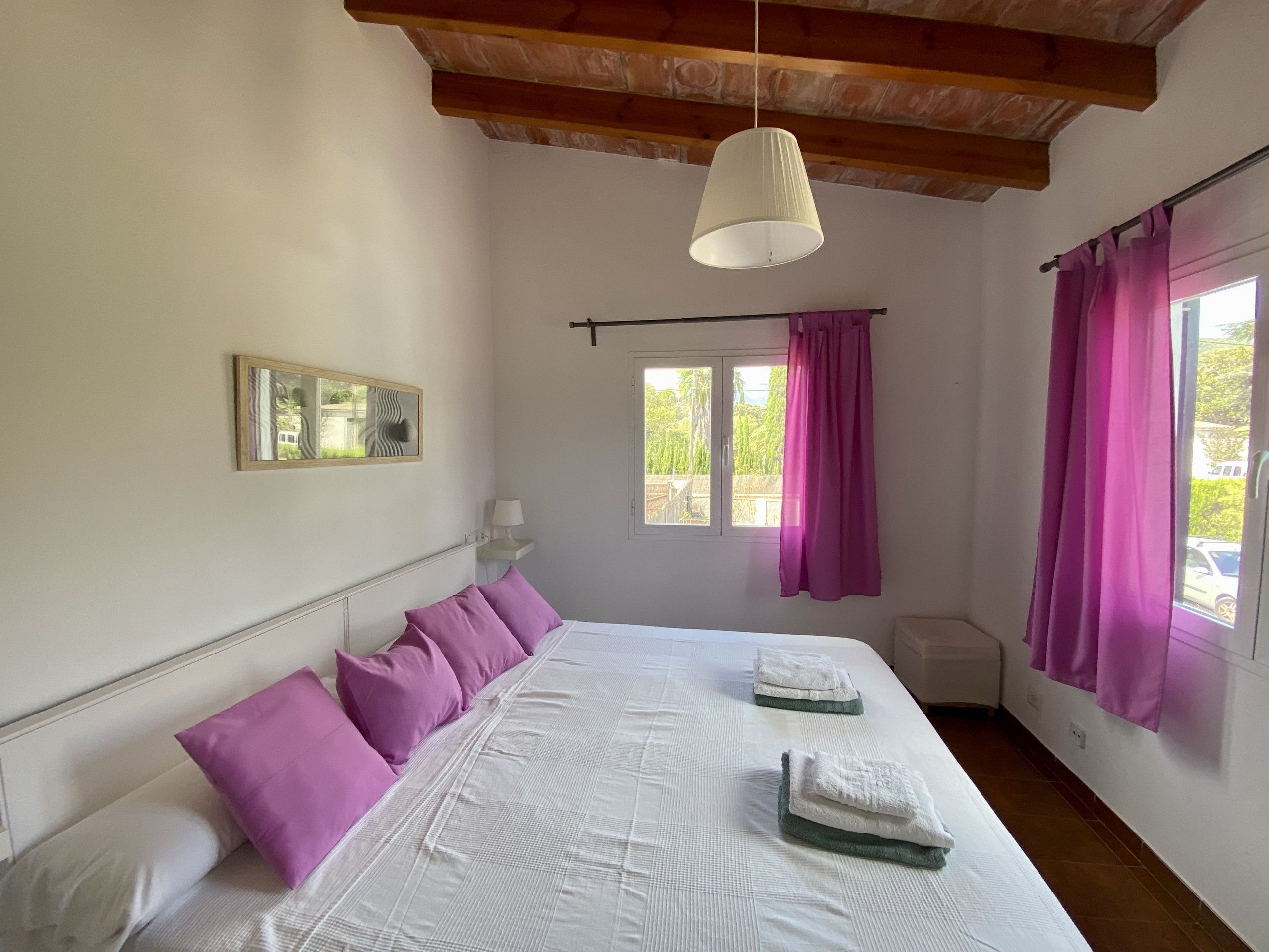 A bedroom with a bed, pillows, a hanging lamp, a window with curtains, and a wall-mounted picture.