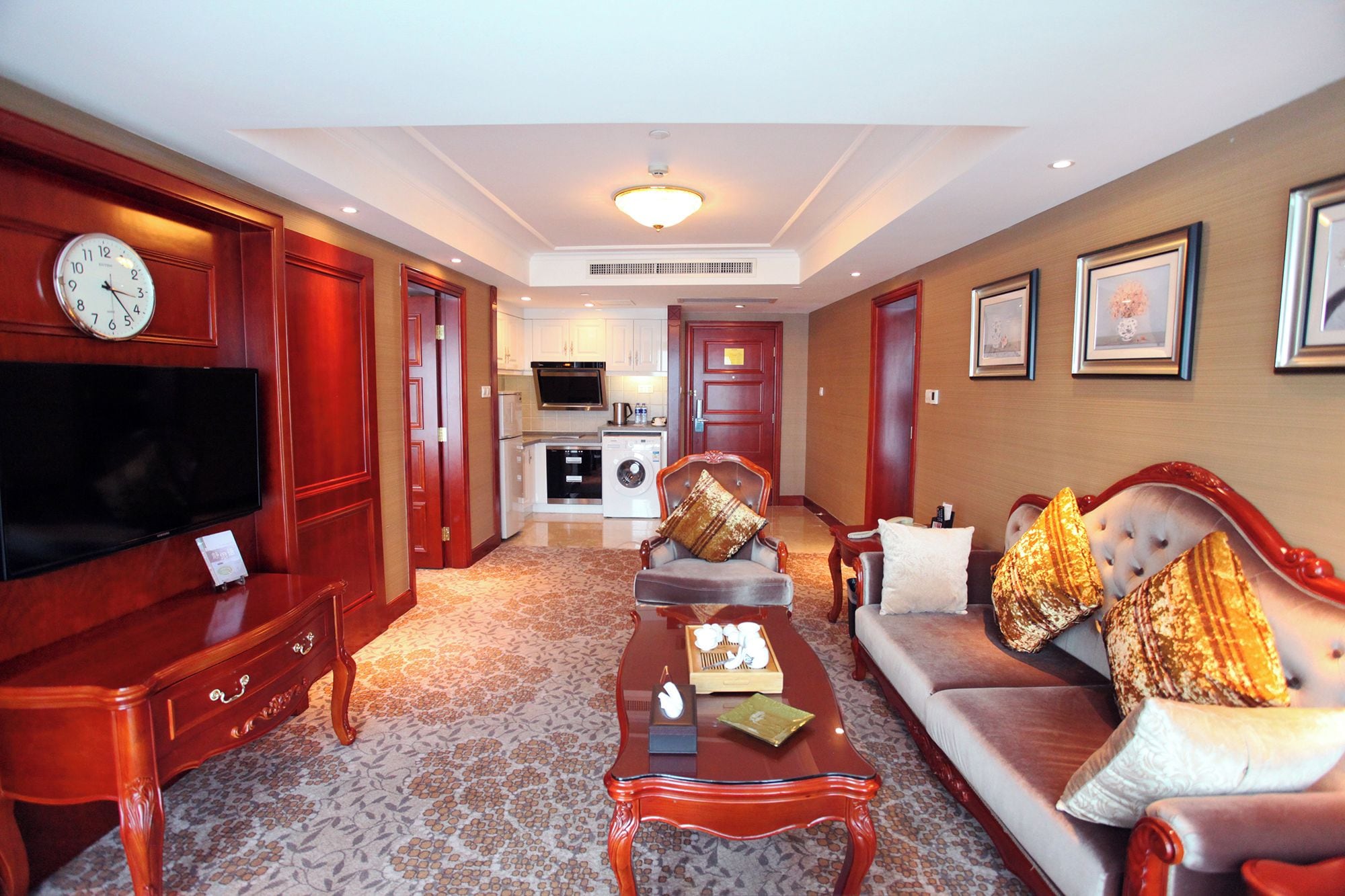 A hotel room with a living area, a sofa set, a television, a coffee table, a clock, and a kitchen area.