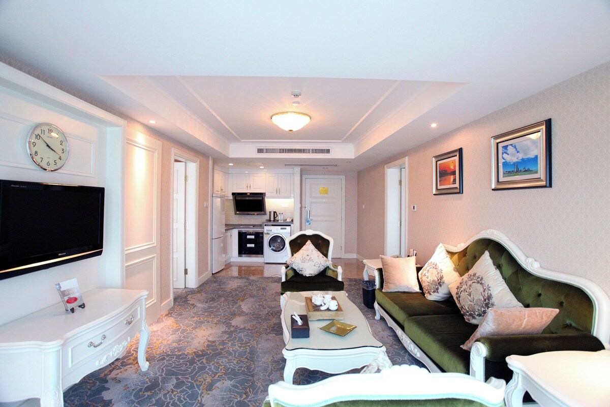 A living room with a flat-screen TV, a sofa, a coffee table, and a dining area with a kitchen in the background.