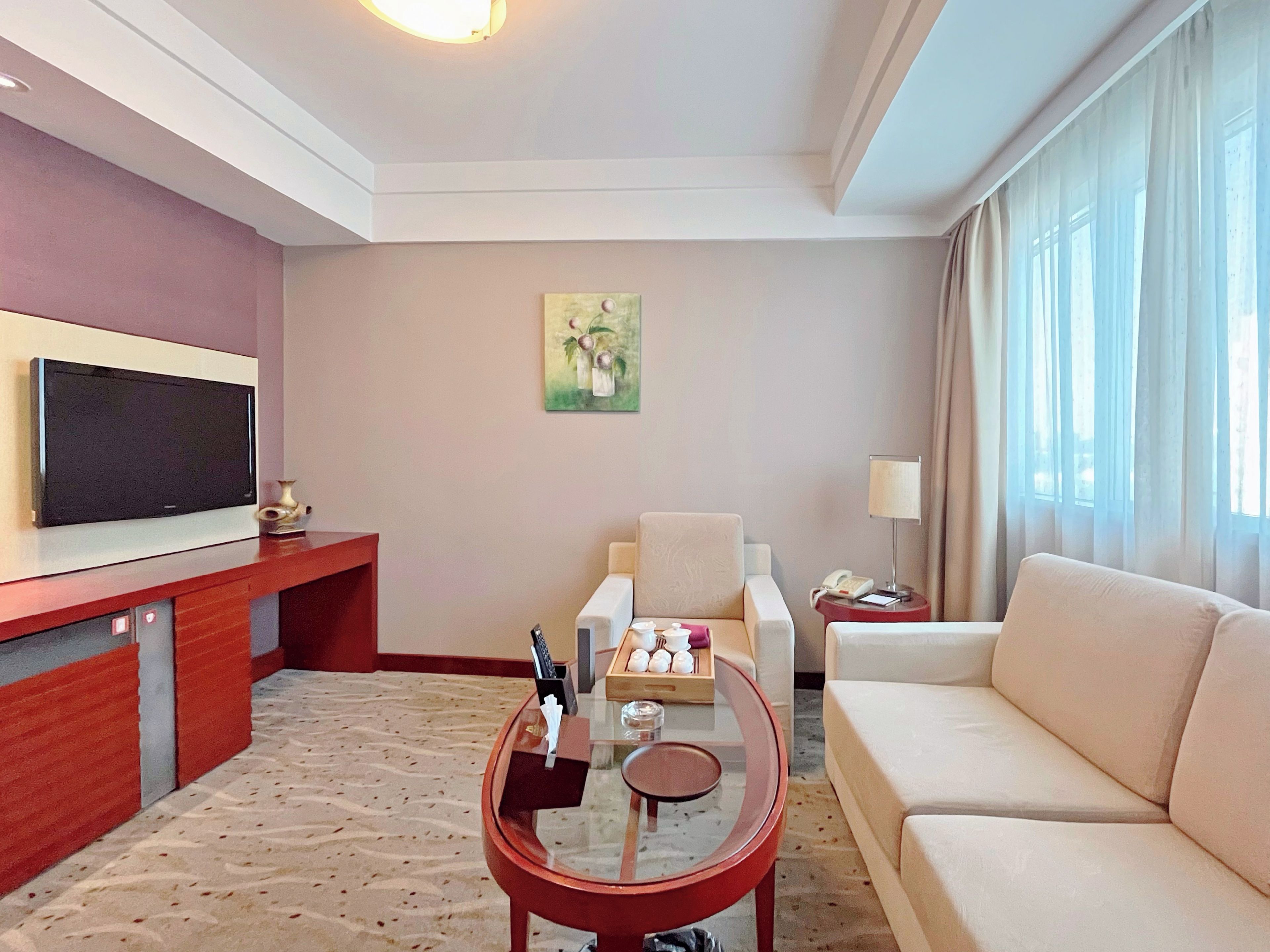 A hotel room with a flat-screen TV, a sofa, a glass coffee table, a painting on the wall, and a window with curtains.