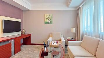 A hotel room with a flat-screen TV, a sofa, a glass coffee table, a painting on the wall, and a window with curtains.
