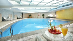 Ein Hallenbad mit Glasdach, Liegestühlen und einem Tisch mit Getränken und Früchten.