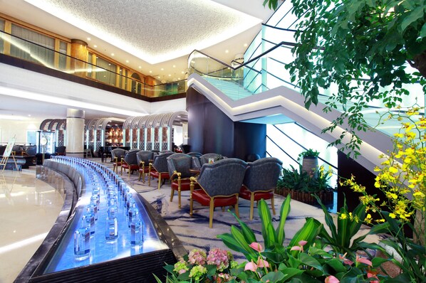 En modern hotellobby med en böjd vattenfontän, en sittgrupp, en trappa och inomhusväxter.