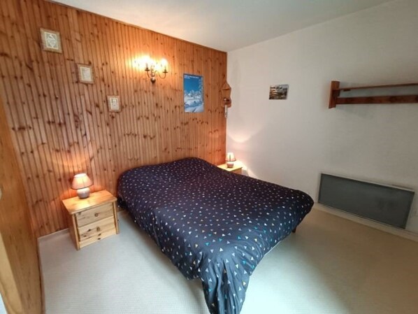 A bedroom with a wooden panel wall, a bed with a dark blue polka dot bedspread, two bedside tables with lamps, and a small window with a view.