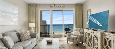 A living room with a sofa, armchairs, a coffee table, and a TV on a wooden cabinet. A sign on the wall reads "Beach House."