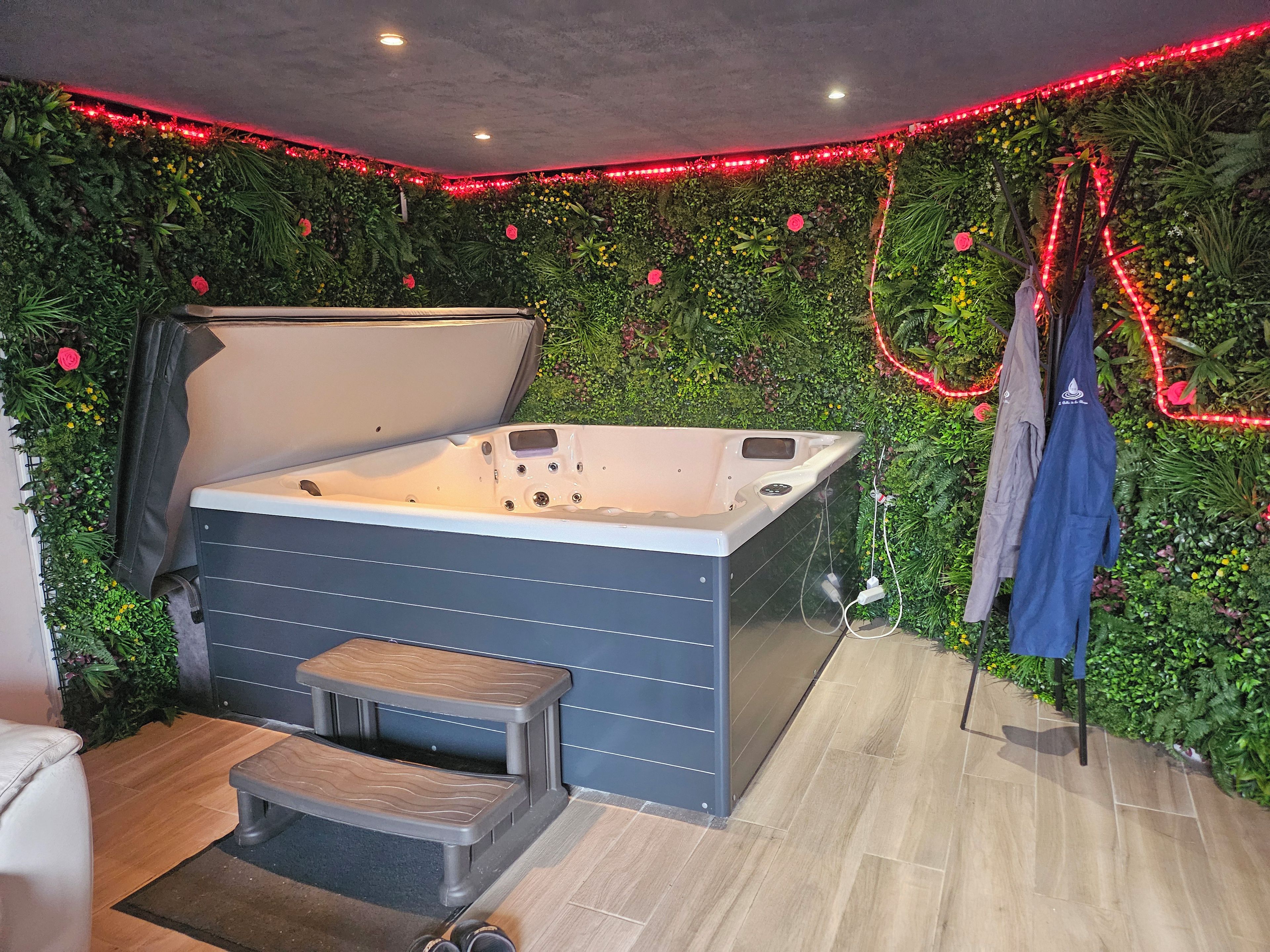 Zona de spa con un jacuzzi, un taburete y una pared cubierta de follaje y luces LED rojas.