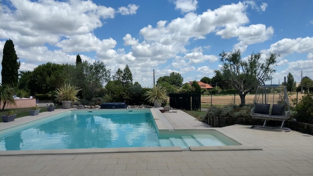 Uma piscina retangular com um deck ladrilhado, rodeada por plantas em vasos e uma rede.